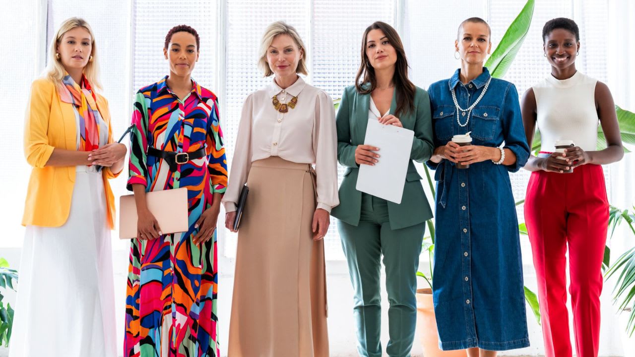 Six women stand indoors, dressed in colorful, professional outfits that perfectly exemplify an executive dress code, holding notebooks and coffee cups as they pose for a group photo against a bright background with plants.